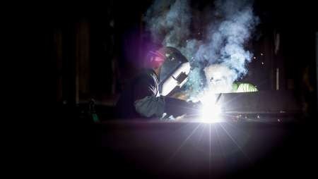 A welder in full mask welding in a darkened workshop with sparks and smoke.