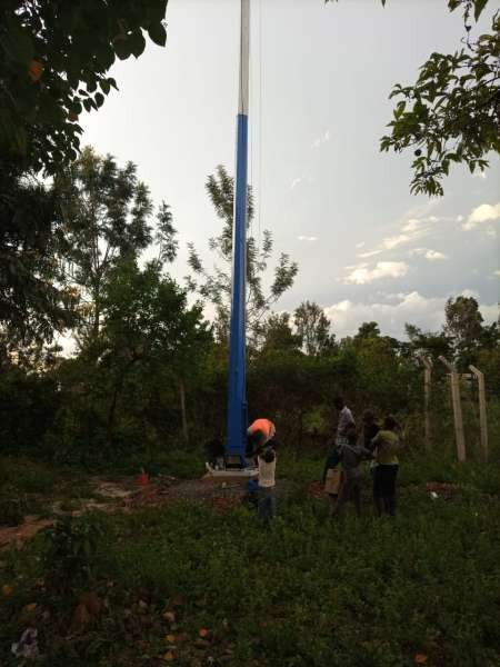 A blue high-mast light pole standing in a rural compound at golden hour with neighbours gathered at the base.