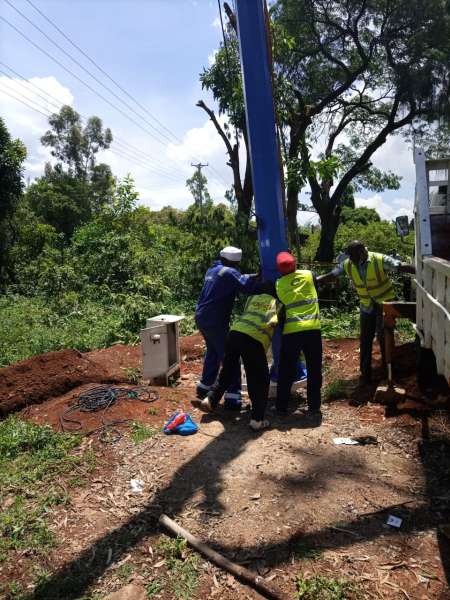 Four installers in hi-vis vests guiding a tall blue light pole into vertical position.