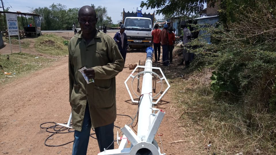 A man in a dark coat poses next to a delivered street-light mast on a dirt road with a Mitsubishi truck behind.