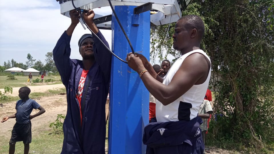 Two Benlight technicians wiring the base of a blue mast while children watch from nearby.