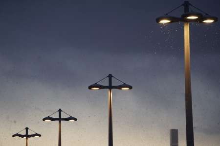 Four street lights coming on at dusk along a rural road.